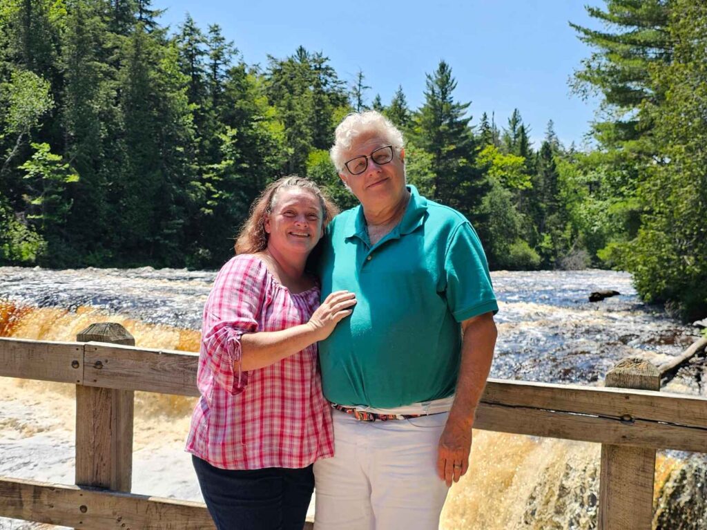 Heather Crosby and husband David standing on a bridge with a small waterfall behind them.