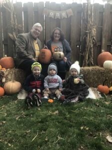 Heather Crosby and husband David with their three young children sitting outside in front of a wooden fence with several pumpkins placed around them.